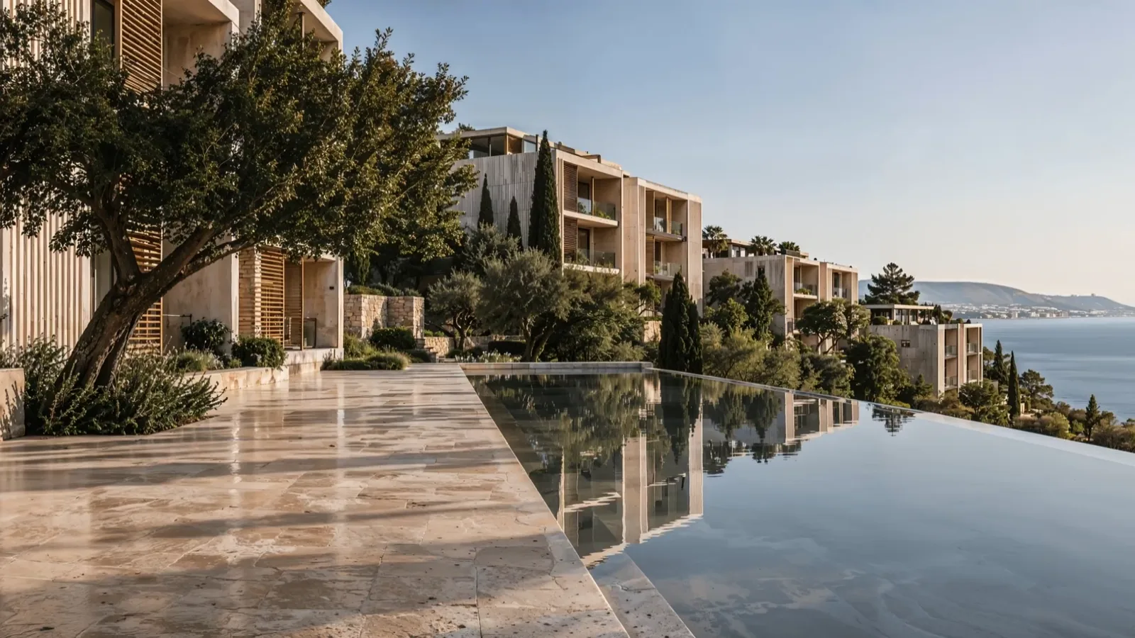 A travertine pool deck beside a long infinity pool, an old olive tree, stone residences stepping down to the sea; the sun sets and the lamps come on. A computer-generated visualisation.