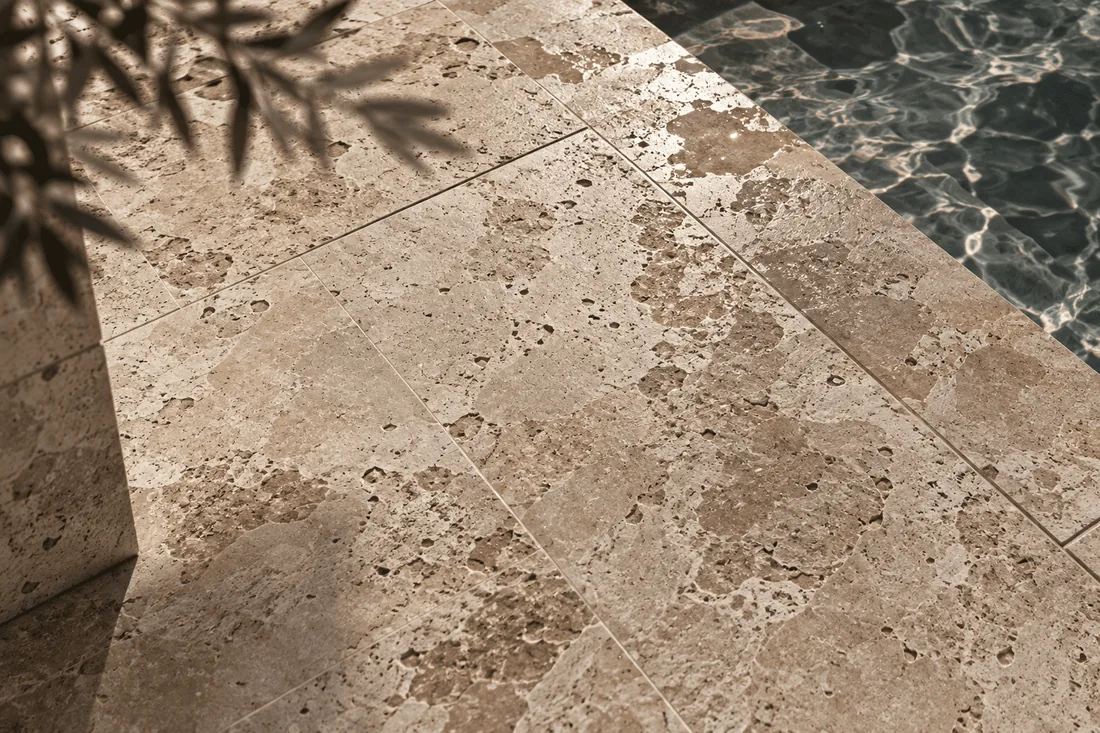 Honed travertine paving at the edge of a pool, olive shadows across it.